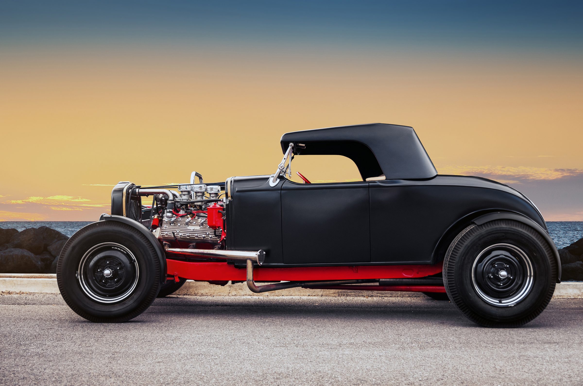 A matte black custom hot rod with a chrome-topped flathead V8 engine, red frame, and whitewall tires, parked at sunset by the ocean.